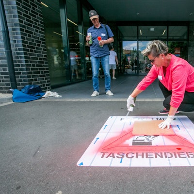 Eine Frau Sprühfarbe auf eine Schablone in Form eines Warndreiecks, deren Schriftzug „Polizei NRW – Taschendiebe“ lautet.