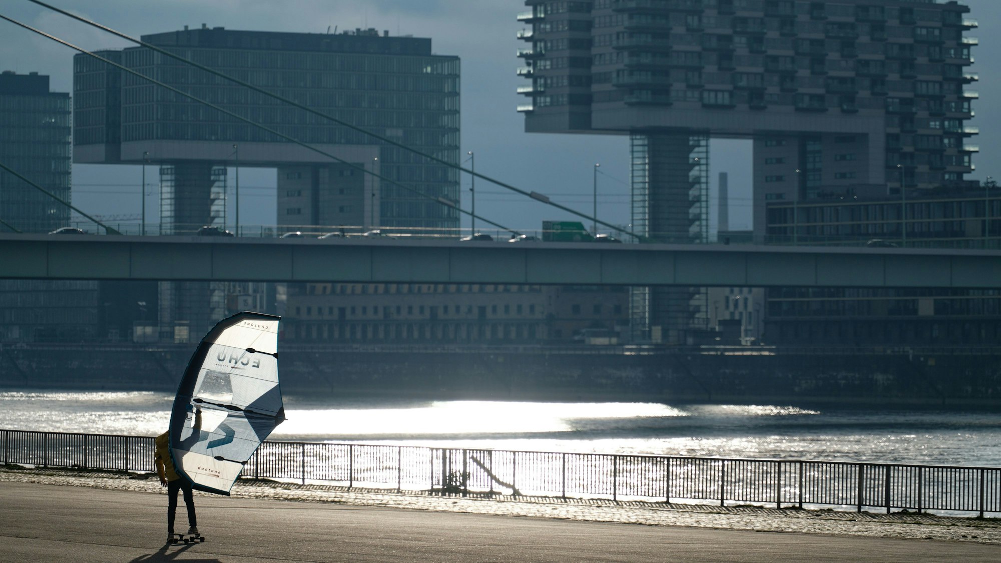 Ein Skateboarder übt sich Freihand im Windskaten mit einem Segel auf einer Freifläche am Rhein.