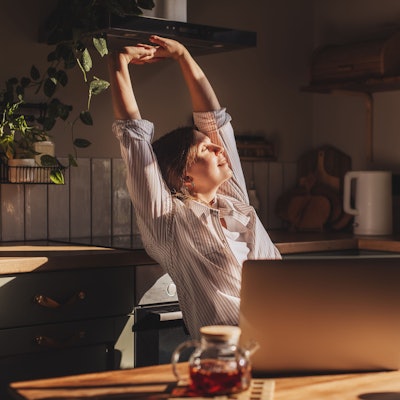 Glückliche, entspannte junge Frau, die in ihrer Küche sitzt, einen Laptop vor sich, die Arme über den Kopf gestreckt und lächelnd aus dem Fenster schaut.