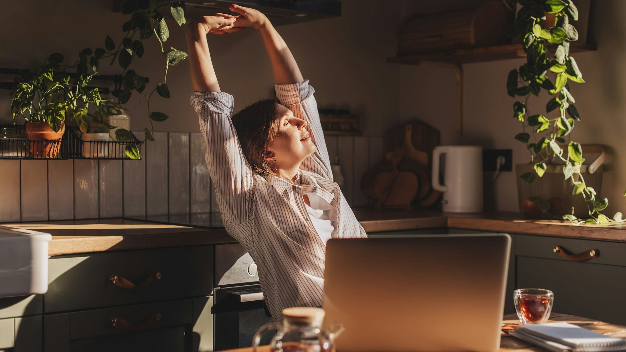 Glückliche, entspannte junge Frau, die in ihrer Küche sitzt, einen Laptop vor sich, die Arme über den Kopf gestreckt und lächelnd aus dem Fenster schaut.