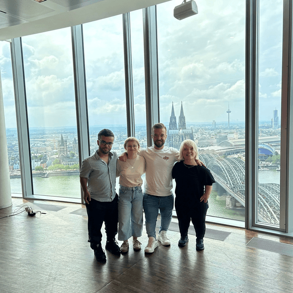 Zu sehen sind vier Menschen. Zwei Frauen und zwei Männer sind zu sehen, die nebeneinander in einem Gebäude mit Glasfassade stehen. Im Hintergrund befindet sich der Kölner Dom.
