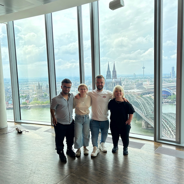 Zu sehen sind vier Menschen. Zwei Frauen und zwei Männer sind zu sehen, die nebeneinander in einem Gebäude mit Glasfassade stehen. Im Hintergrund befindet sich der Kölner Dom.
