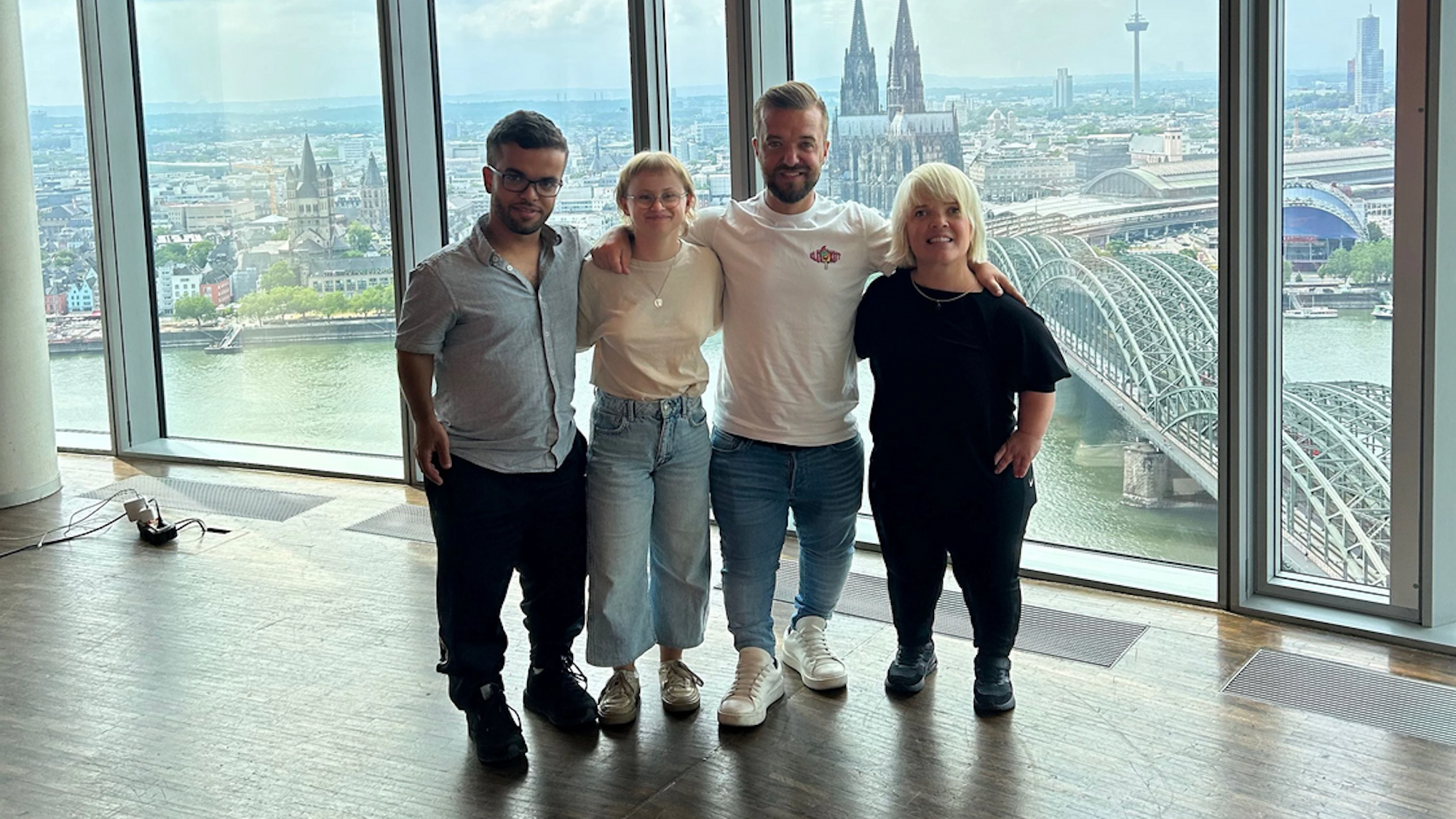 Zu sehen sind vier Menschen. Zwei Frauen und zwei Männer sind zu sehen, die nebeneinander in einem Gebäude mit Glasfassade stehen. Im Hintergrund befindet sich der Kölner Dom.