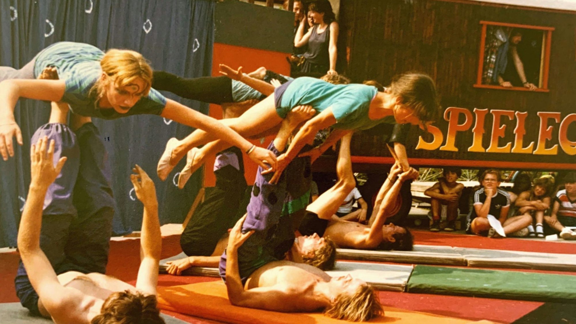 Vier jugendliche Männer liegen auf dem Boden und jonglieren Mädchen auf ihren Füßen.