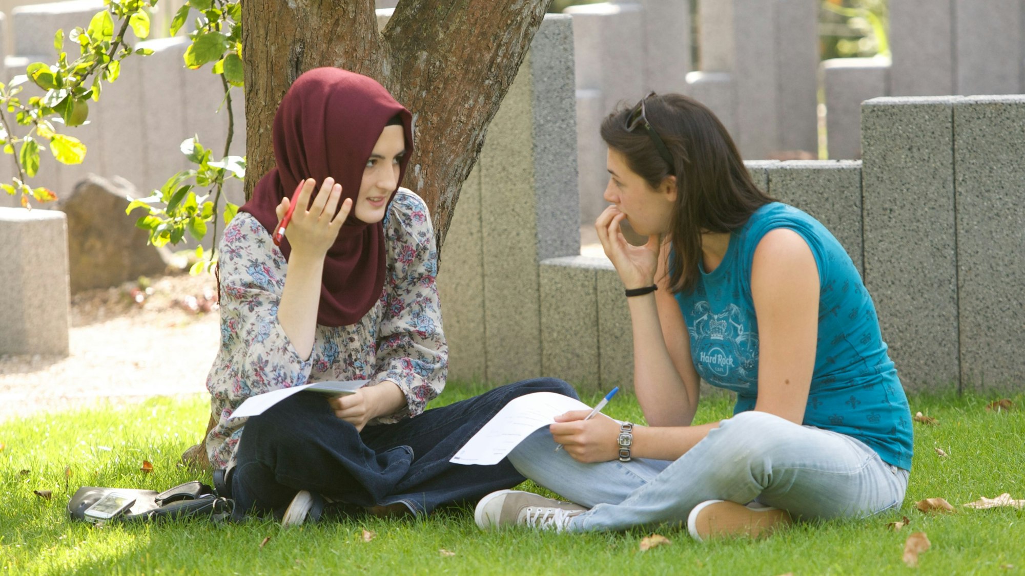 Zwei jugendliche Frauen, eine trägt ein Kopftuch, sitzen im Kölner Garten der Religionen und unterhalten sich.