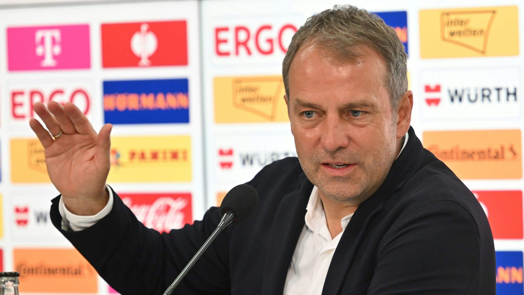 Fußballbundestrainer Hansi Flick spricht nach der 0:2-Niederlage der deutschen Nationalelf gegen Kolumbien auf einer Pressekonferenz. Er sitzt vor einer Wand, auf der Namen von Sponsoren abgebildet sind. Seine rechte Hand ist zur Unterstützung seiner Ausführungen. Er wirkt niedergeschlagen.