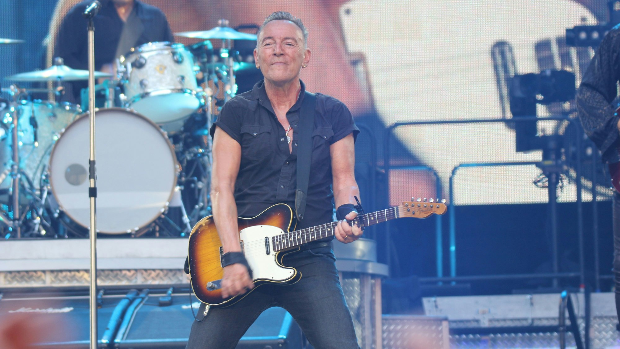 Bruce Springsteen trägt eine schwarze Hose und ein schwarzes kurzärmeliges Hemd. Er steht breitbeinig mit Gitarre vor dem Podest seines Schlagzeugers Max Weinberg.