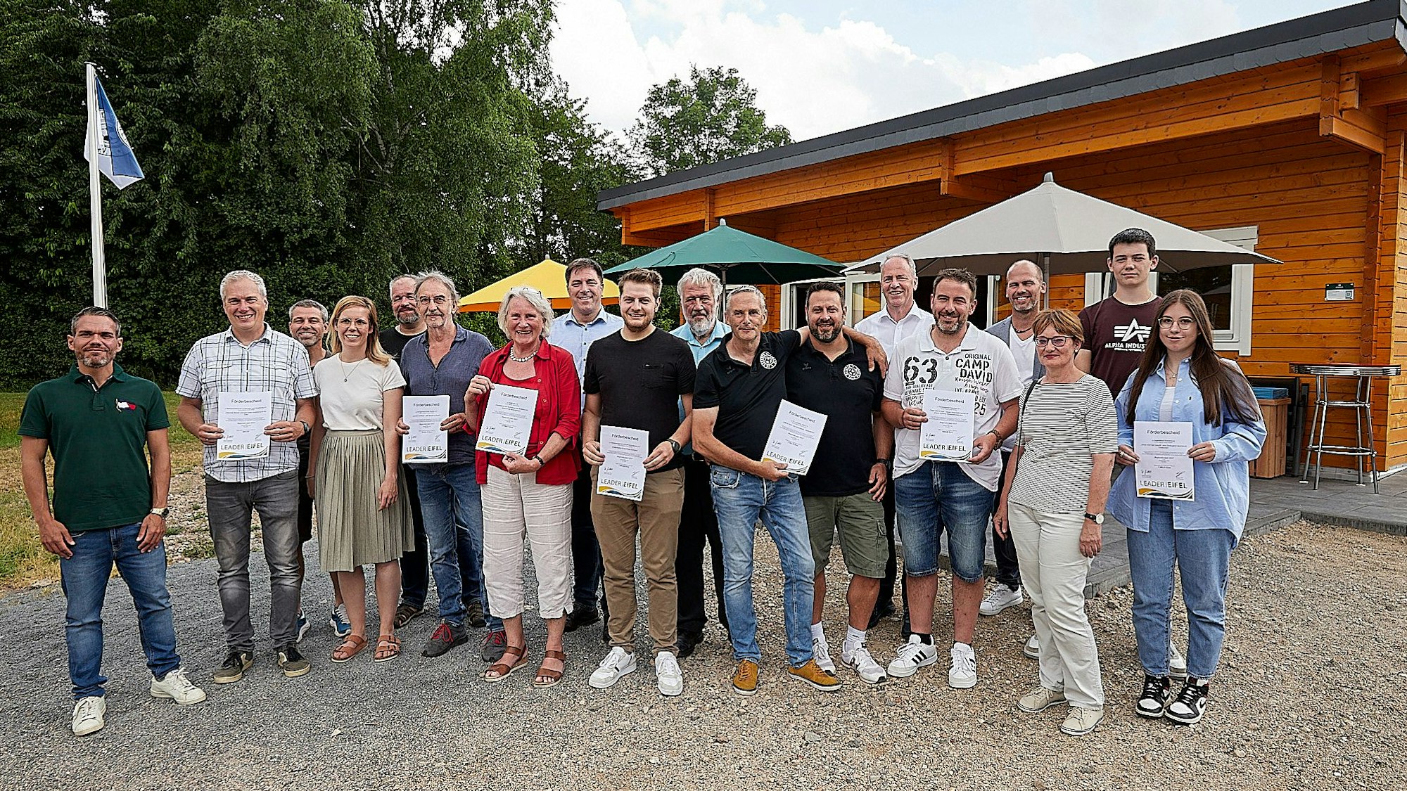 Eine Gruppe von Männern und Frauen halten Förderbescheide in der Hand. Sie stehen vor einem Holzhaus.