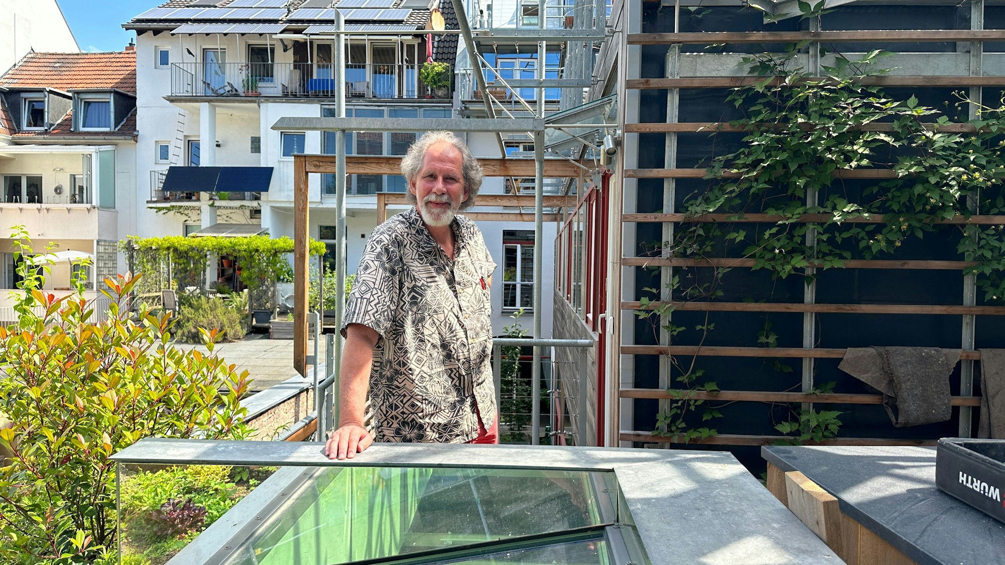 Ein Mann mit grauem Haar und Bart steht im kurzärmeligen Hemd auf einer Dachterrasse.