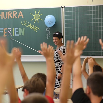 „Hurra Sommerferien“ steht an einer Tafel in der zweiten Klasse einer Grundschule. Eine Lehrerin zeigt mit einem Zeigestock lächelnd auf die Worte, die Kinder heben jubelnd ihre Arme.