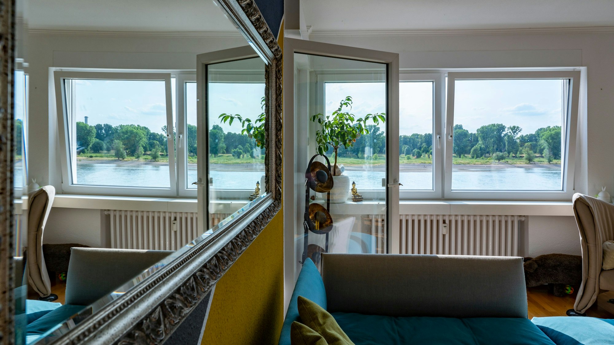 Wohnzimmer der Familie Zosel: Durch zwei Fenster blickt man auf den Rhein und das andere Rheinufer mit Wiese und Bäumen.
