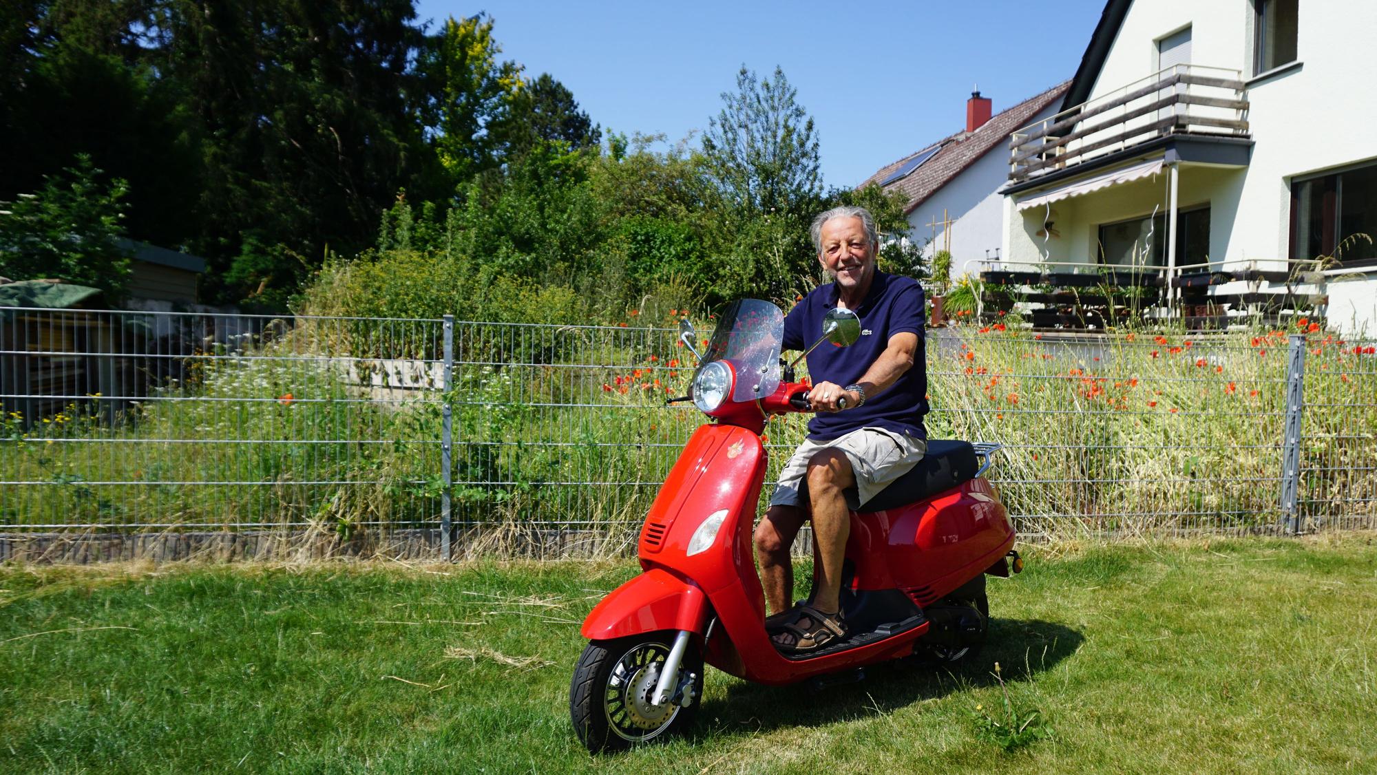Ein 75-jähriger Mann sitzt auf einem roten Roller. Der Roller befindet sich auf der Wiese eines Gartens. Im Hintergrund befinden sich Wohnhäuser.
