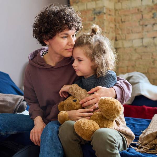 Eine junge Mutter mit gelocktem Kurzhaarschnitt hält ihre kleine Tochter im Arm, die einen Teddybären festhält.