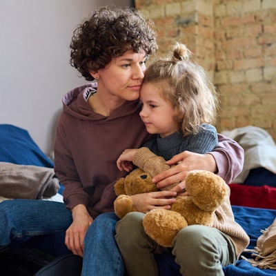 Eine junge Mutter mit gelocktem Kurzhaarschnitt hält ihre kleine Tochter im Arm, die einen Teddybären festhält.