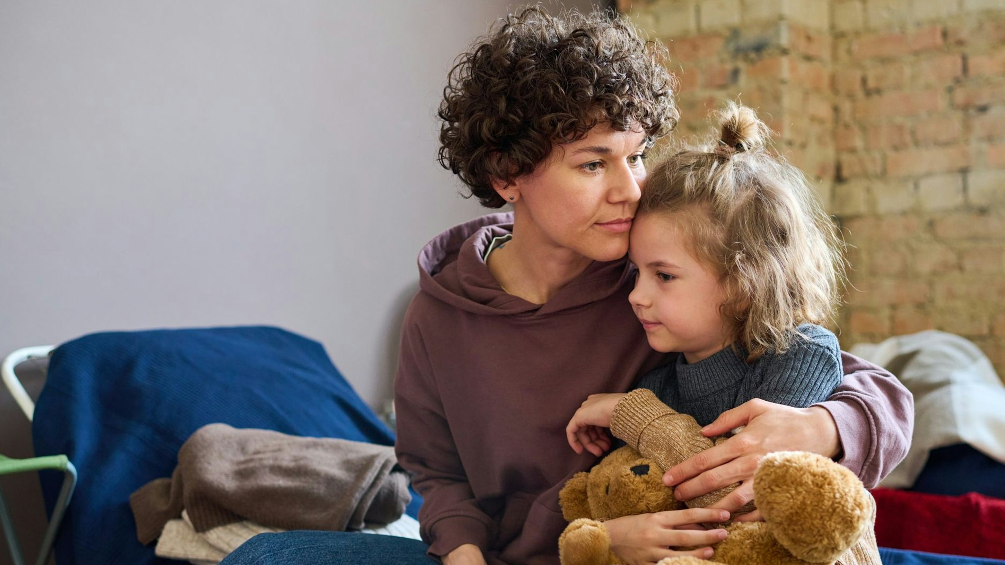 Eine junge Mutter mit gelocktem Kurzhaarschnitt hält ihre kleine Tochter im Arm, die einen Teddybären festhält.