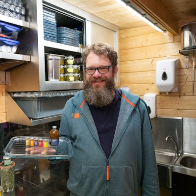 Ein Mann mit langem Bart und Brille hält ein Tablett in der Hand, auf dem kleine Fläschen mit bunten Gewürzen stehen.