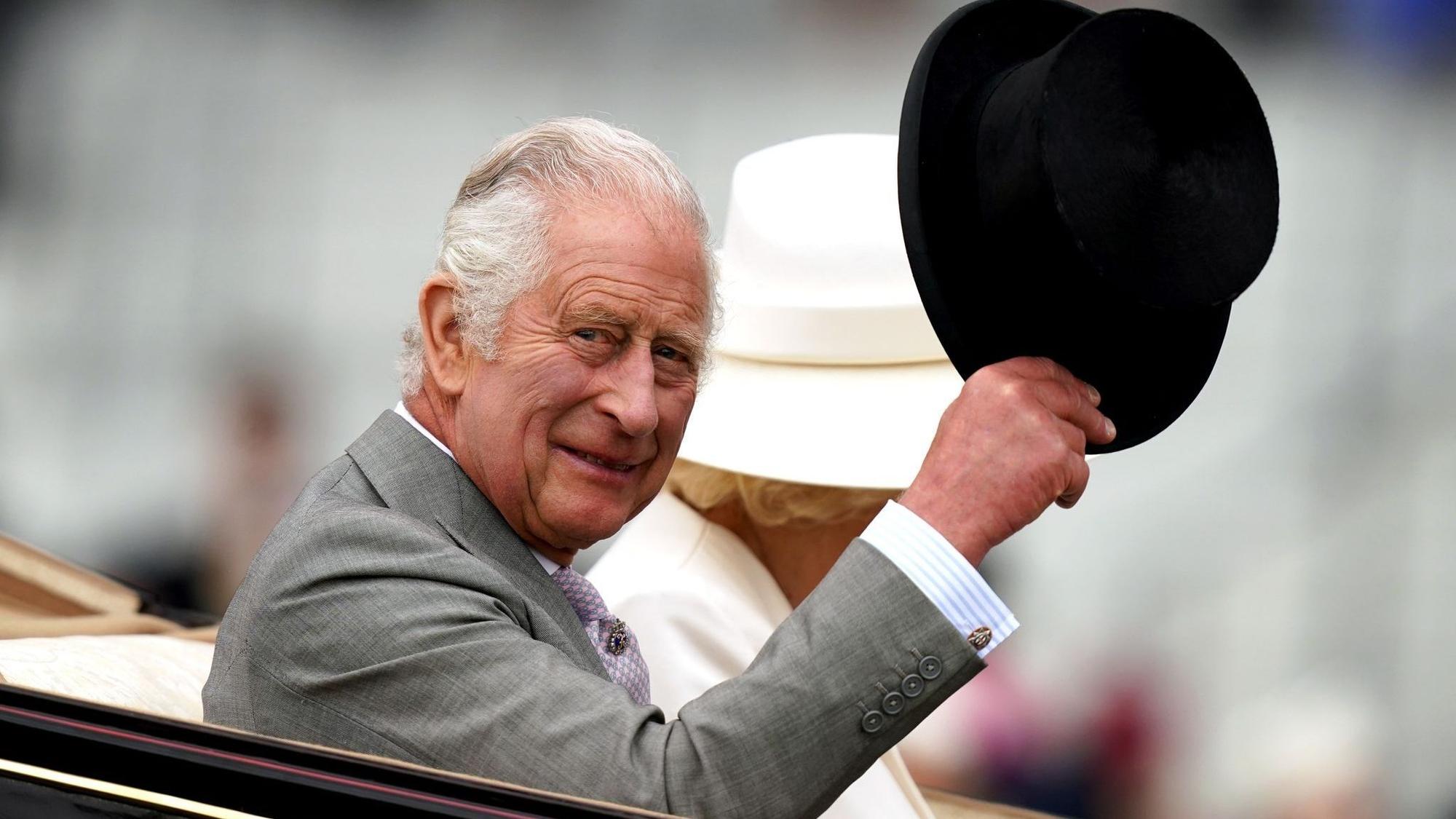 König Charles III. hebt seinen Hut. Neben ihm sitzt Camilla in der Kutsche.