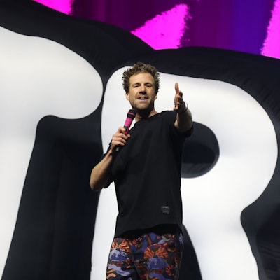 Luke Mockridge steht in einem schwarzen T-Shirt, einer bunten Hose und mit einem pinken Mikrofon in der Kölner Lanxess-Arena auf der Bühne.