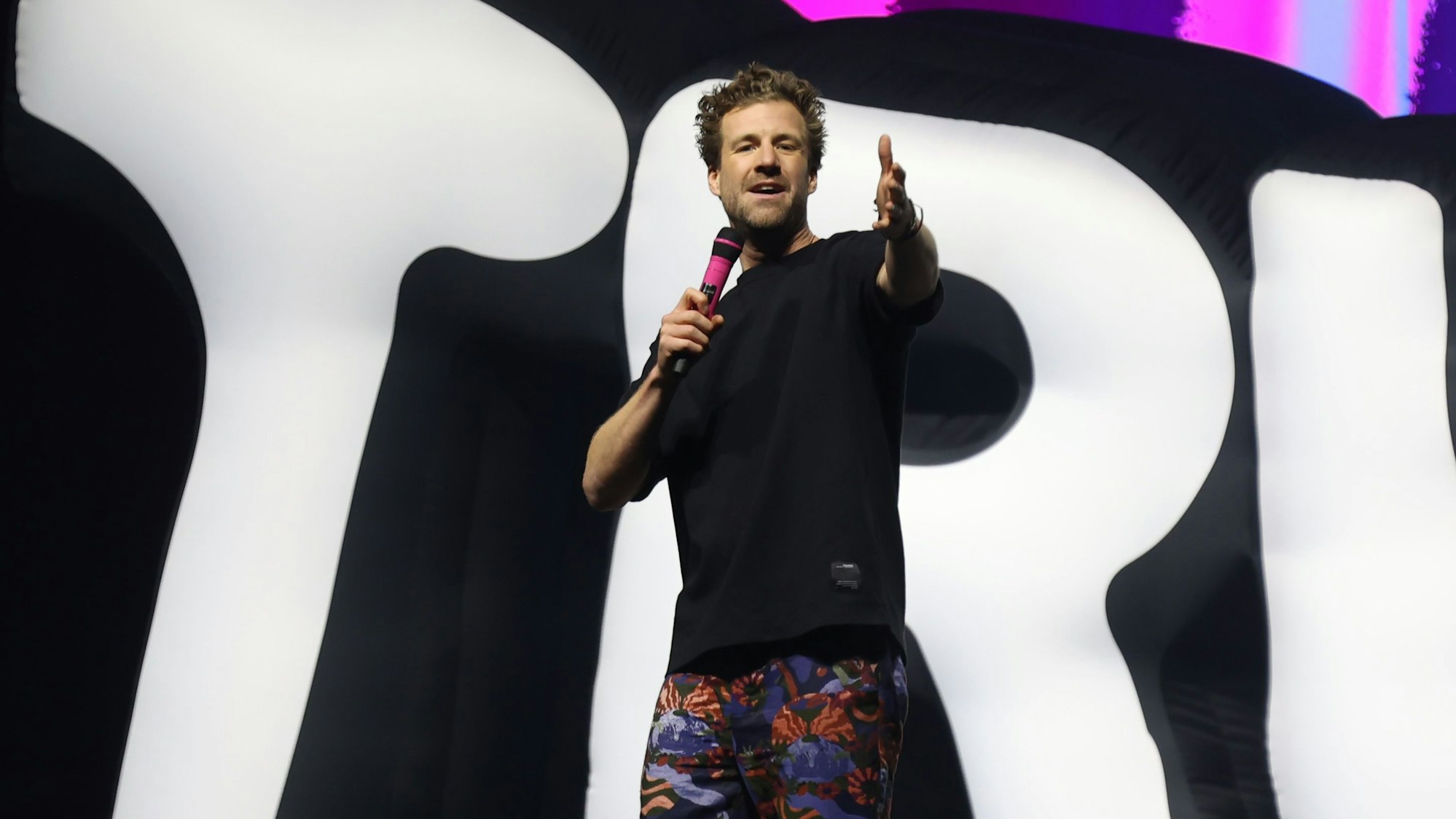 Luke Mockridge steht in einem schwarzen T-Shirt, einer bunten Hose und mit einem pinken Mikrofon in der Kölner Lanxess-Arena auf der Bühne.