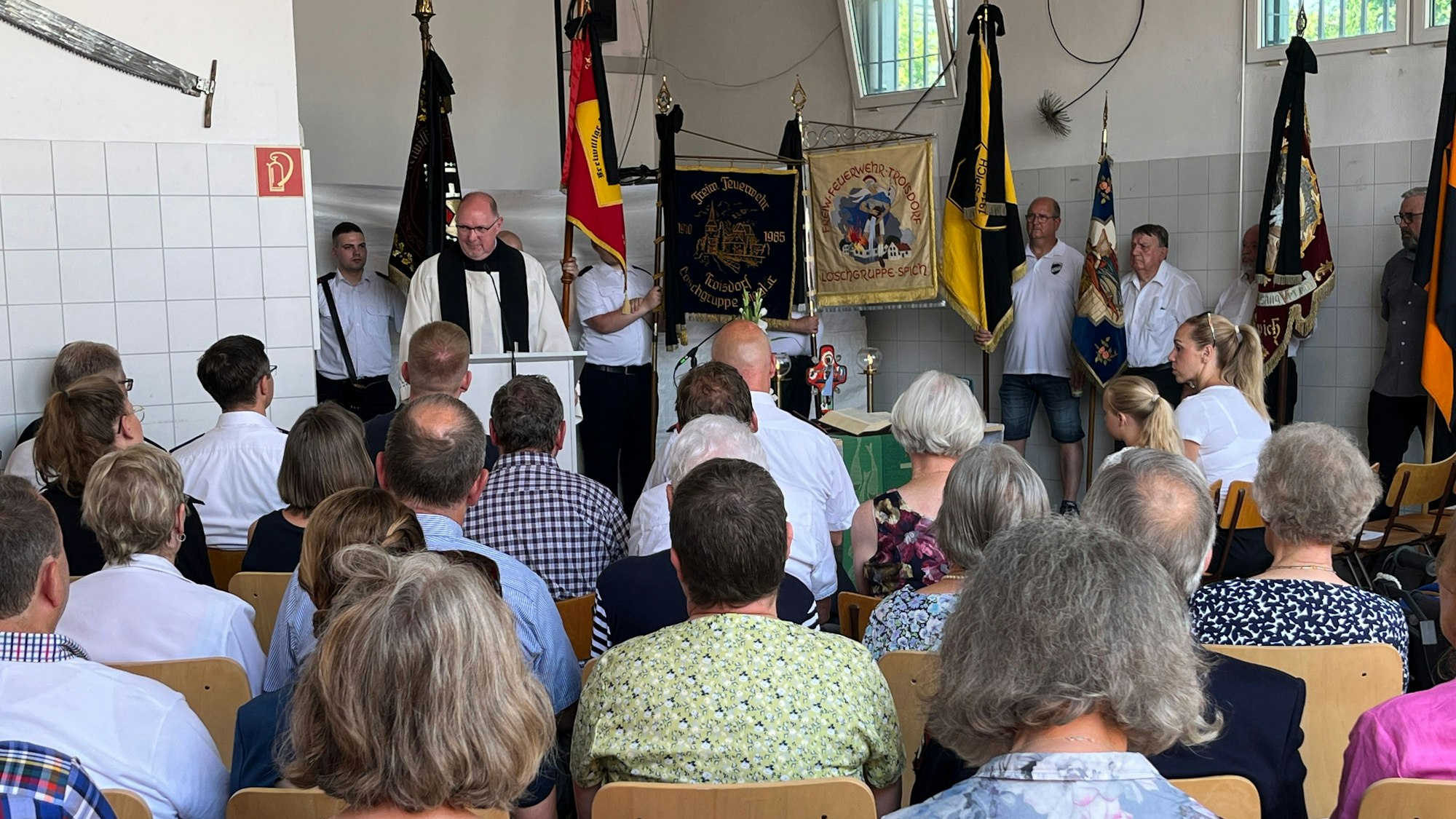 Im Gerätehaus spricht Pfarrer Hermann-Josef Zeyen vor den Flaggen der Einheiten stehend zu den Besucherinnen und Besuchern.