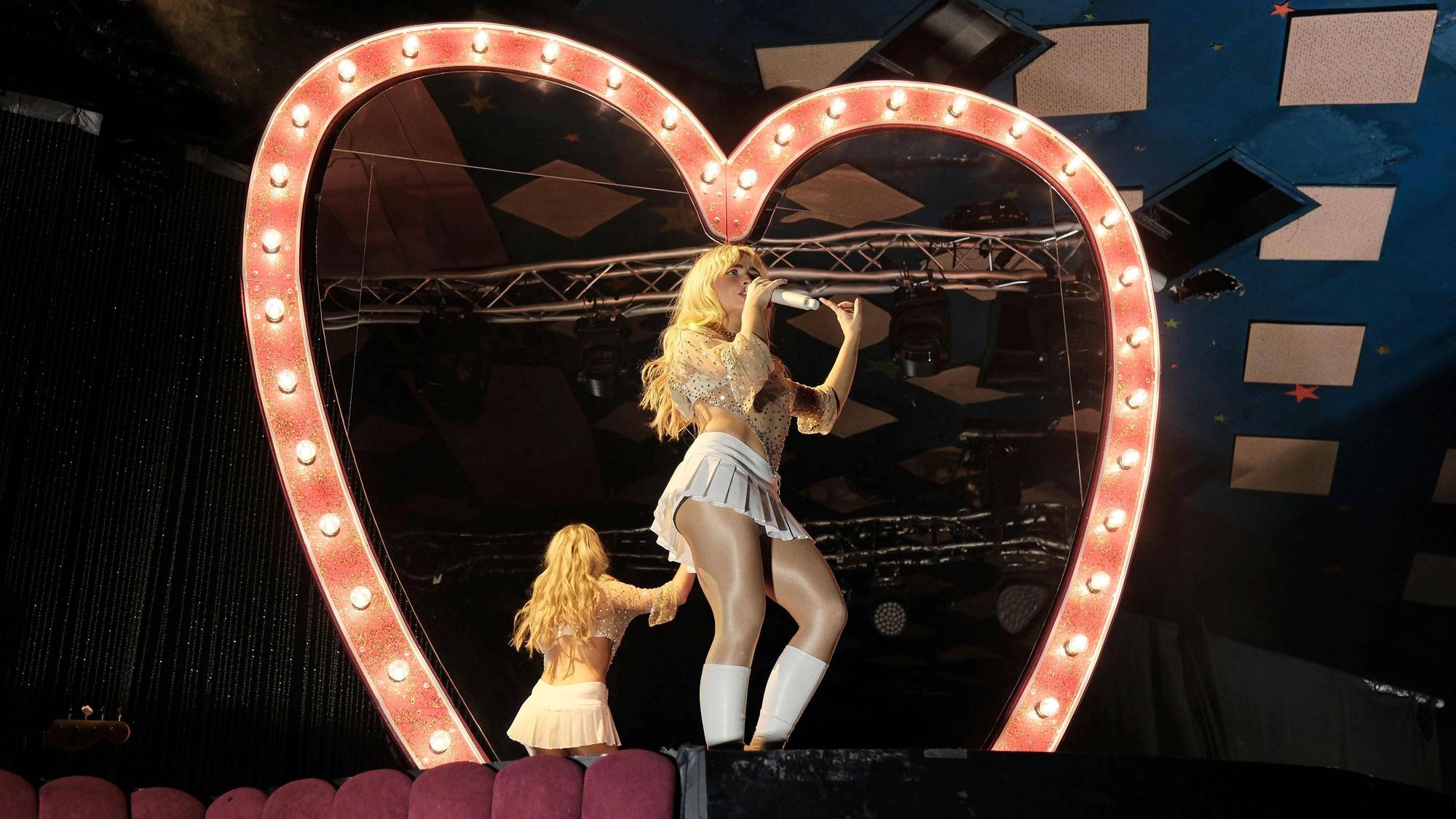 Sabrina Carpenter steht in weißem Minirock mit dem Rücken zu einem großen herzförmigen Spiegel.