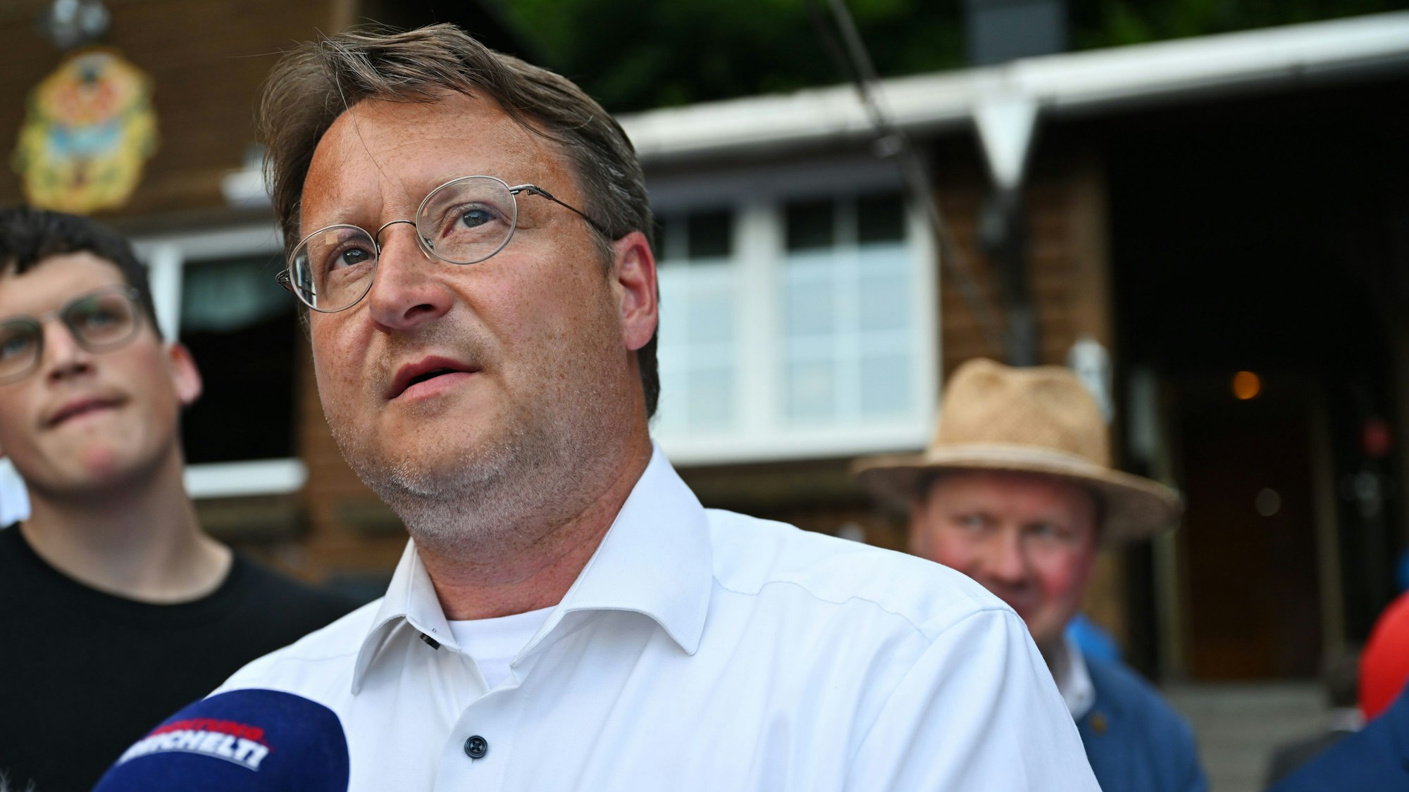 Robert Sesselmann trägt ein weißes Hemd. Er steht vor einem Mikrofon und gibt ein Interview.