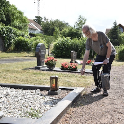 Ein älterer Mann deutet auf ein Grab auf einem Friedhof.