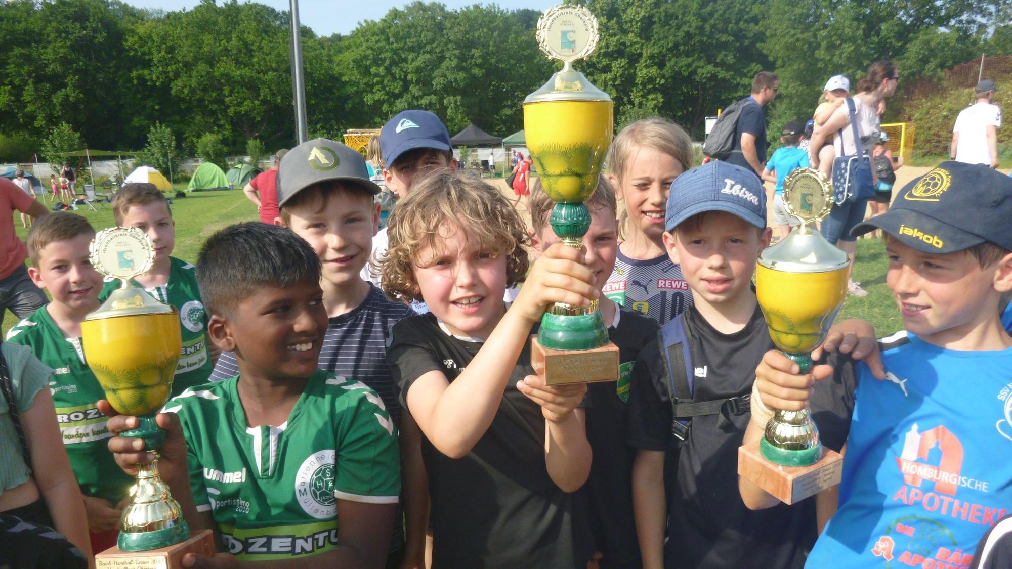Mehrere Jungs halten nach einem Beachhandballturnier stolz die gewonnenen Pokale in die Höhe.