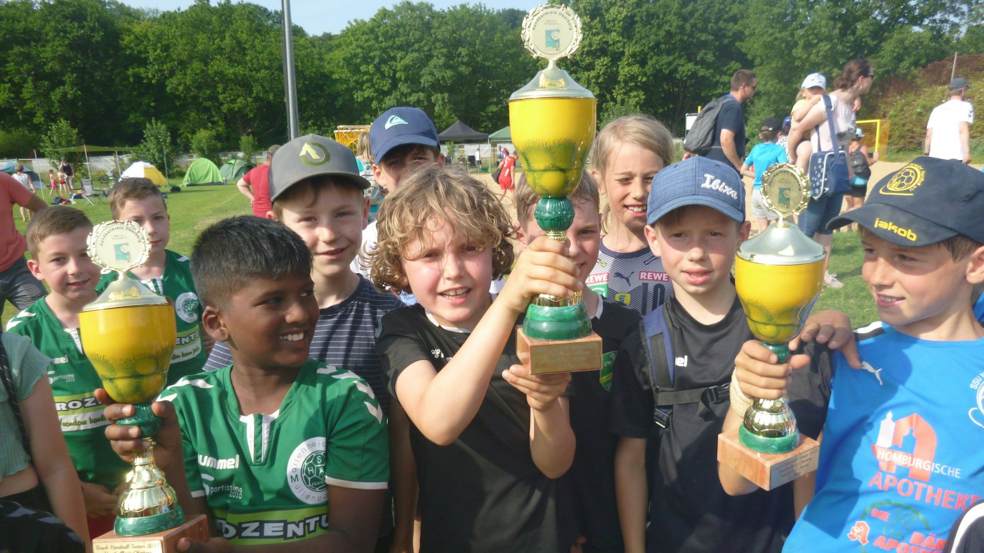 Mehrere Jungs halten nach einem Beachhandballturnier stolz die gewonnenen Pokale in die Höhe.