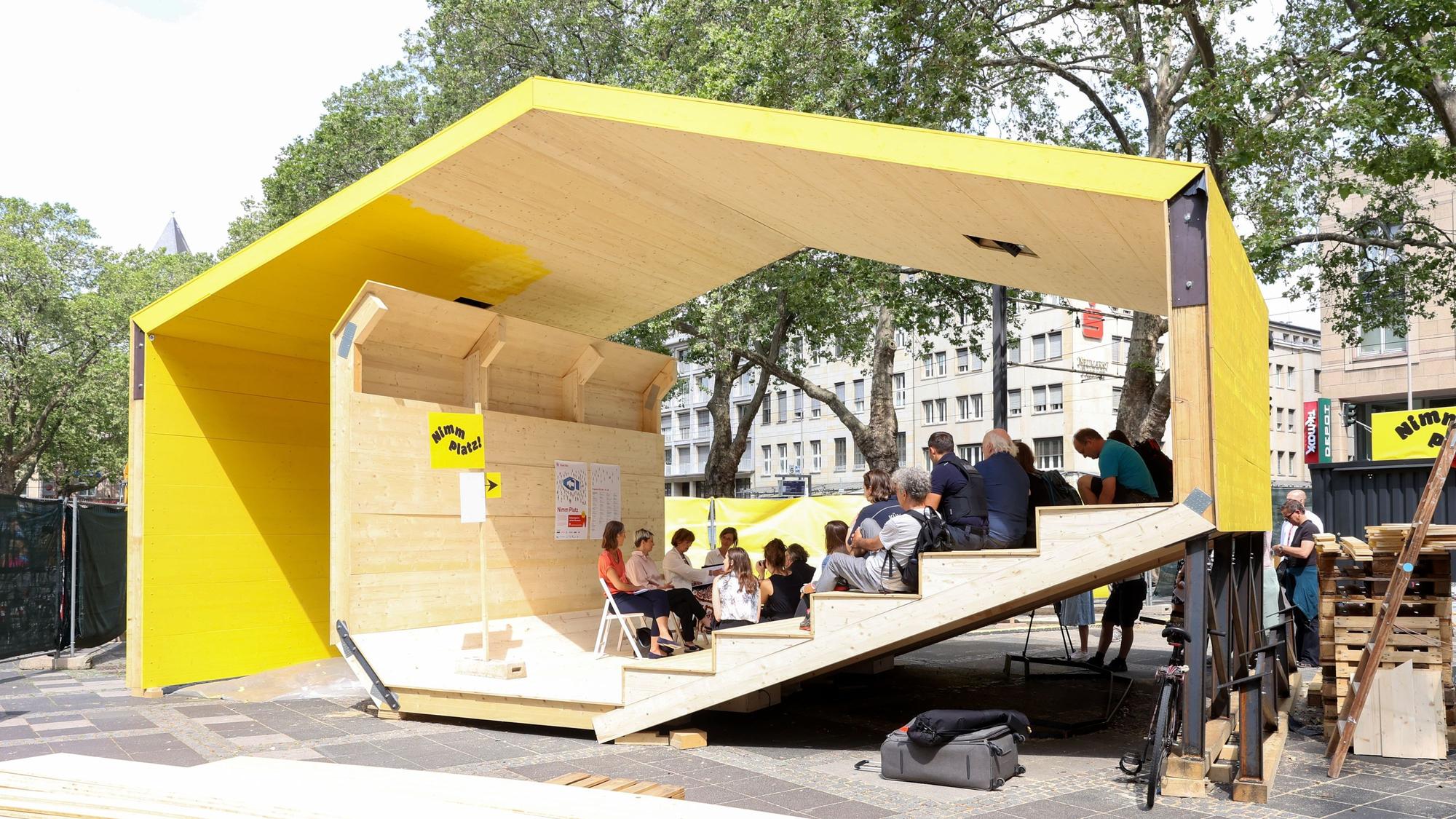 Ein Holzpavillon in gelber Farbe auf einem Platz.