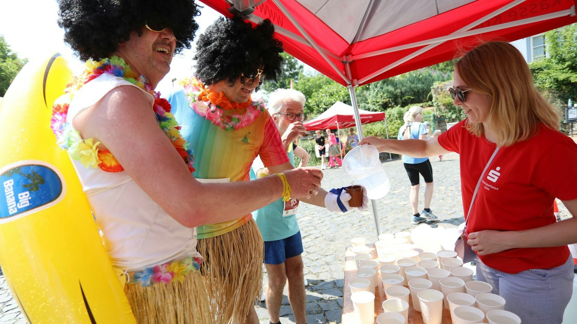 Zwei Männer mit Baströcken und schwarzen Perücke lassen sich an einem Getränkestand mit Wasser versorgen. Auf einem Holztisch stehen gefüllte Becher.