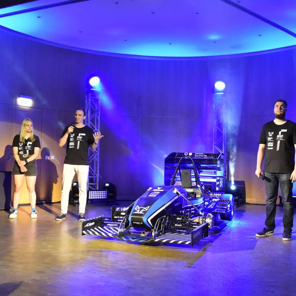 Rollout des neuen Rennwagens der Hochschule Bonn-Rhein-Sieg: Experten erklären die neu entwickelten Bauteile. Drei Männer und eine Frau stehen neben dem Rennwagen auf der Bühne.