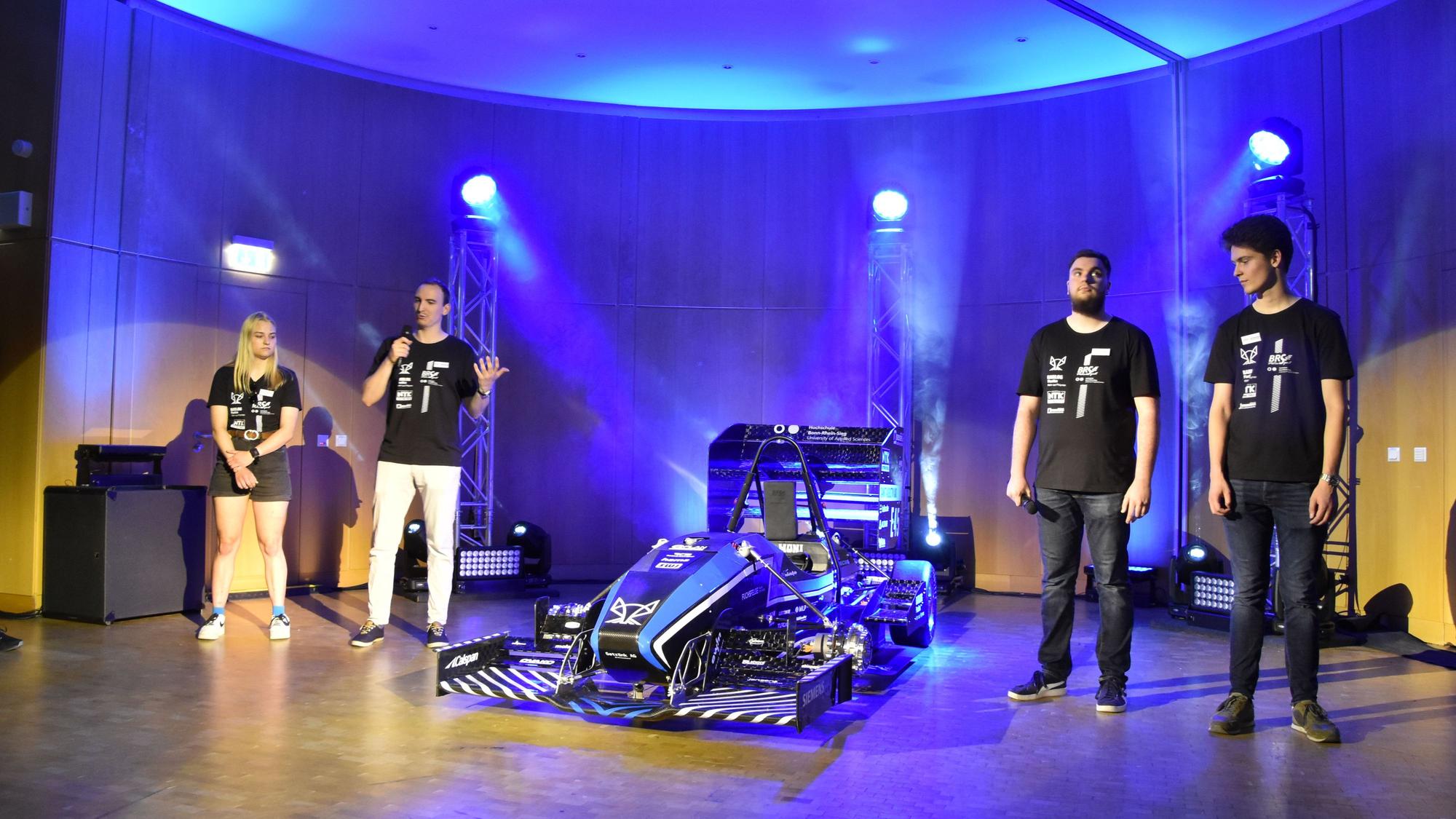 Rollout des neuen Rennwagens der Hochschule Bonn-Rhein-Sieg: Experten erklären die neu entwickelten Bauteile. Drei Männer und eine Frau stehen neben dem Rennwagen auf der Bühne.