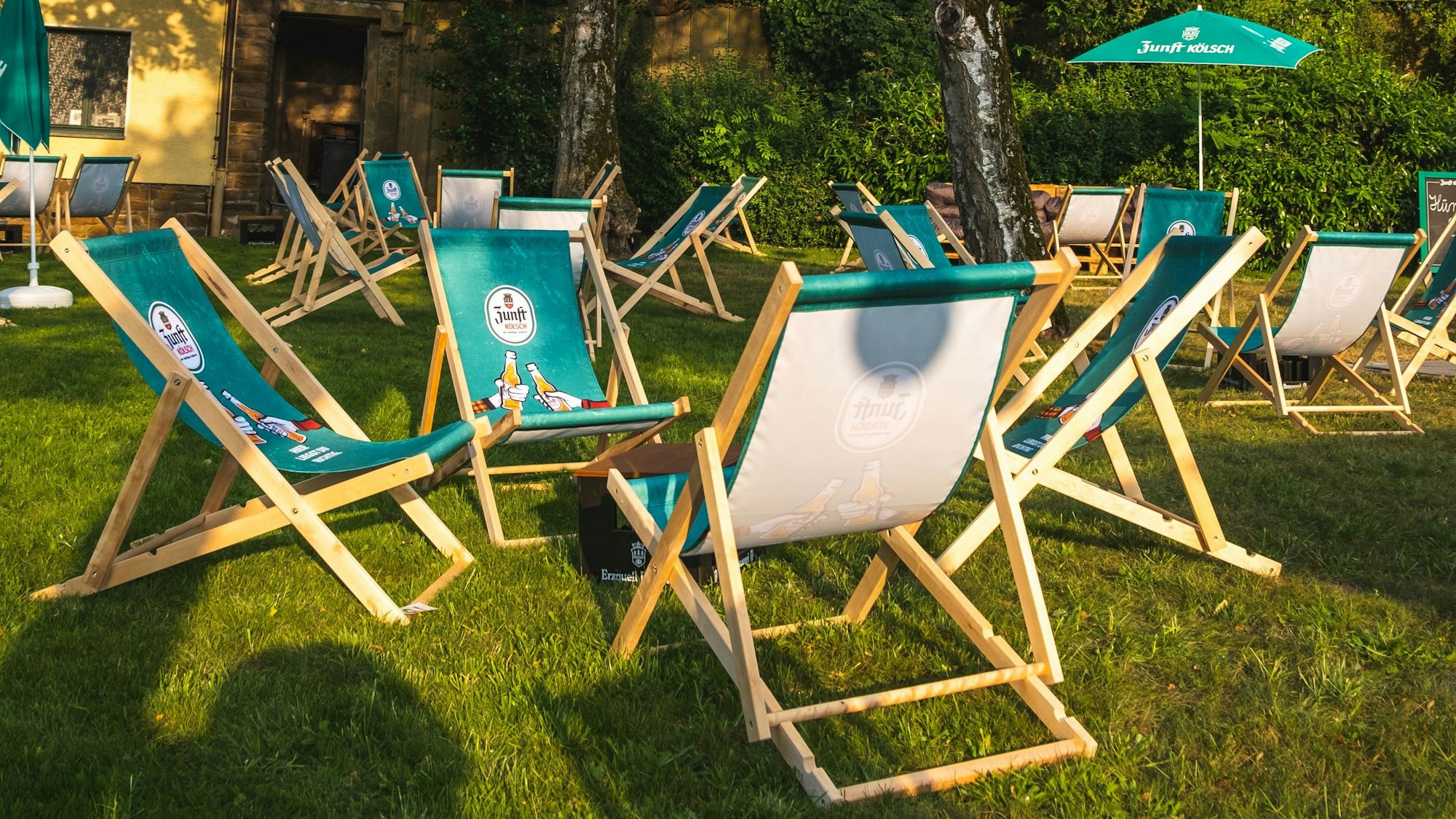 Auf einer Wiese stehen mehrere grüne Liegestühle mit Zunft Kölsch Logo.