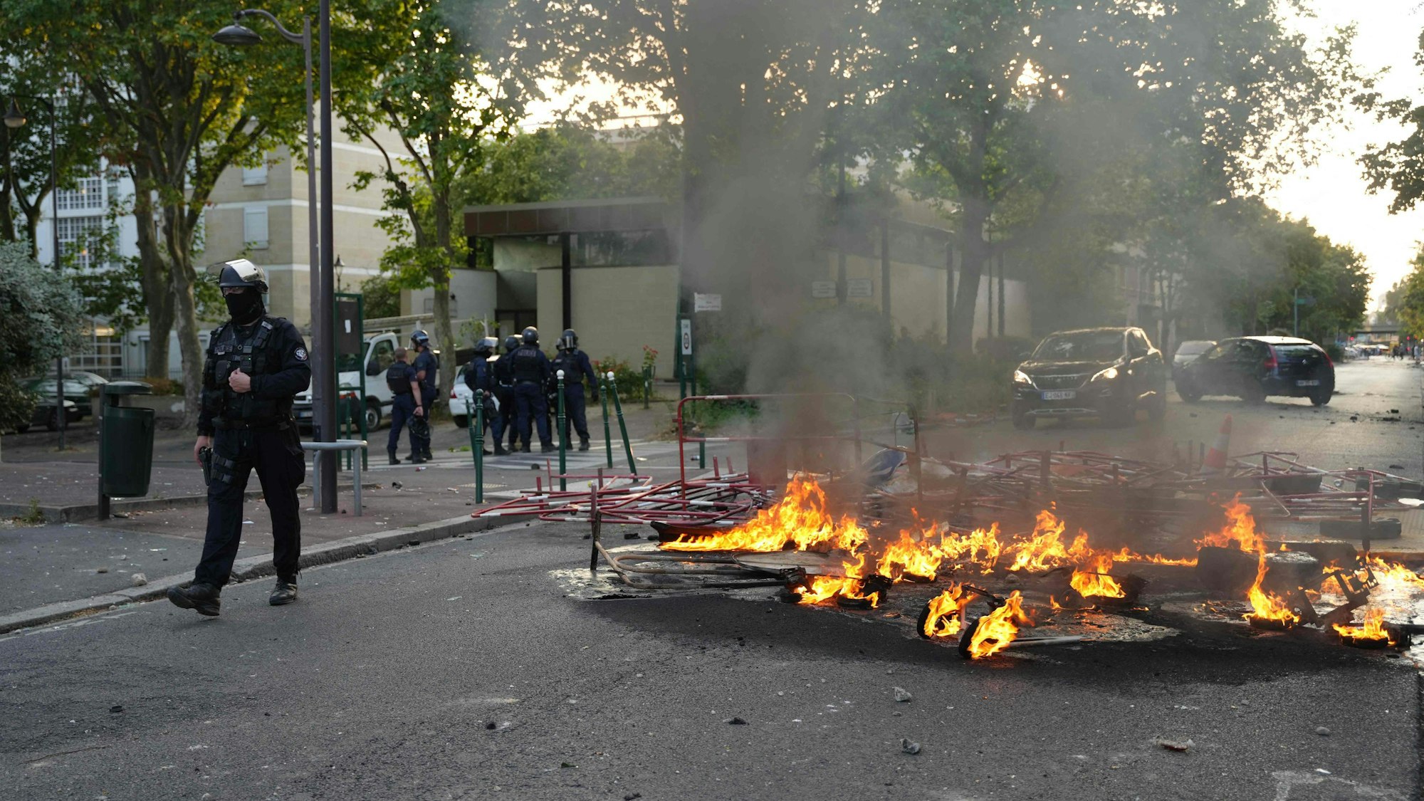Polizisten in Einsatzkleidung stehen neben einem Feuer, das nach einer Demonstration in Nanterre, westlich von Paris, am 27. Juni 2023 auf der Straße brennt, nachdem die französische Polizei einen Jugendlichen erschossen hatte, der sich geweigert hatte, bei einer Verkehrskontrolle in der Stadt anzuhalten.