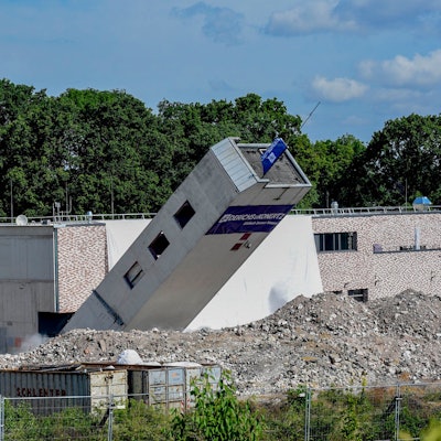 Das Foto zeigt einen umkippenden Turm vor einem Neubau.