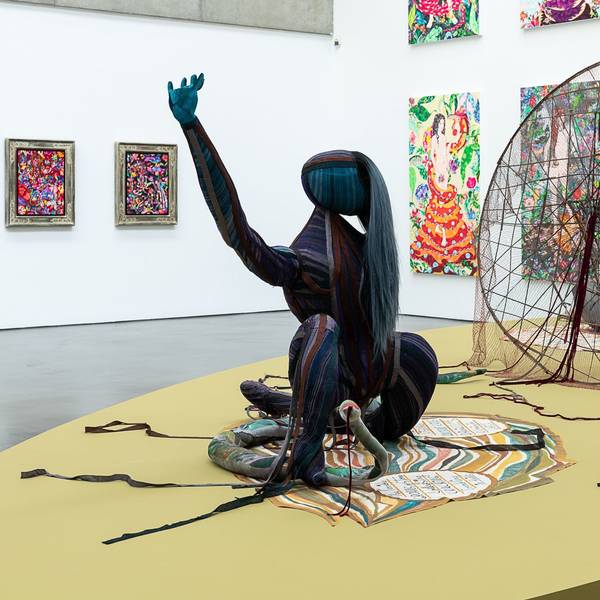 Im Vordergrund ist eine Skulptur auf einem Teppich, dahinter ein großes traumfängerartiges Netz und einige Bilder an den Wänden.