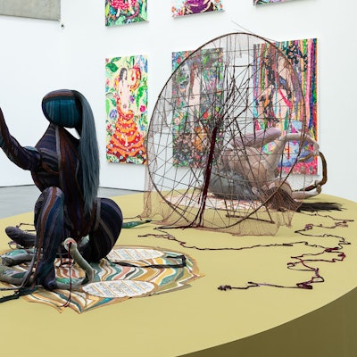 Im Vordergrund ist eine Skulptur auf einem Teppich, dahinter ein großes traumfängerartiges Netz und einige Bilder an den Wänden.