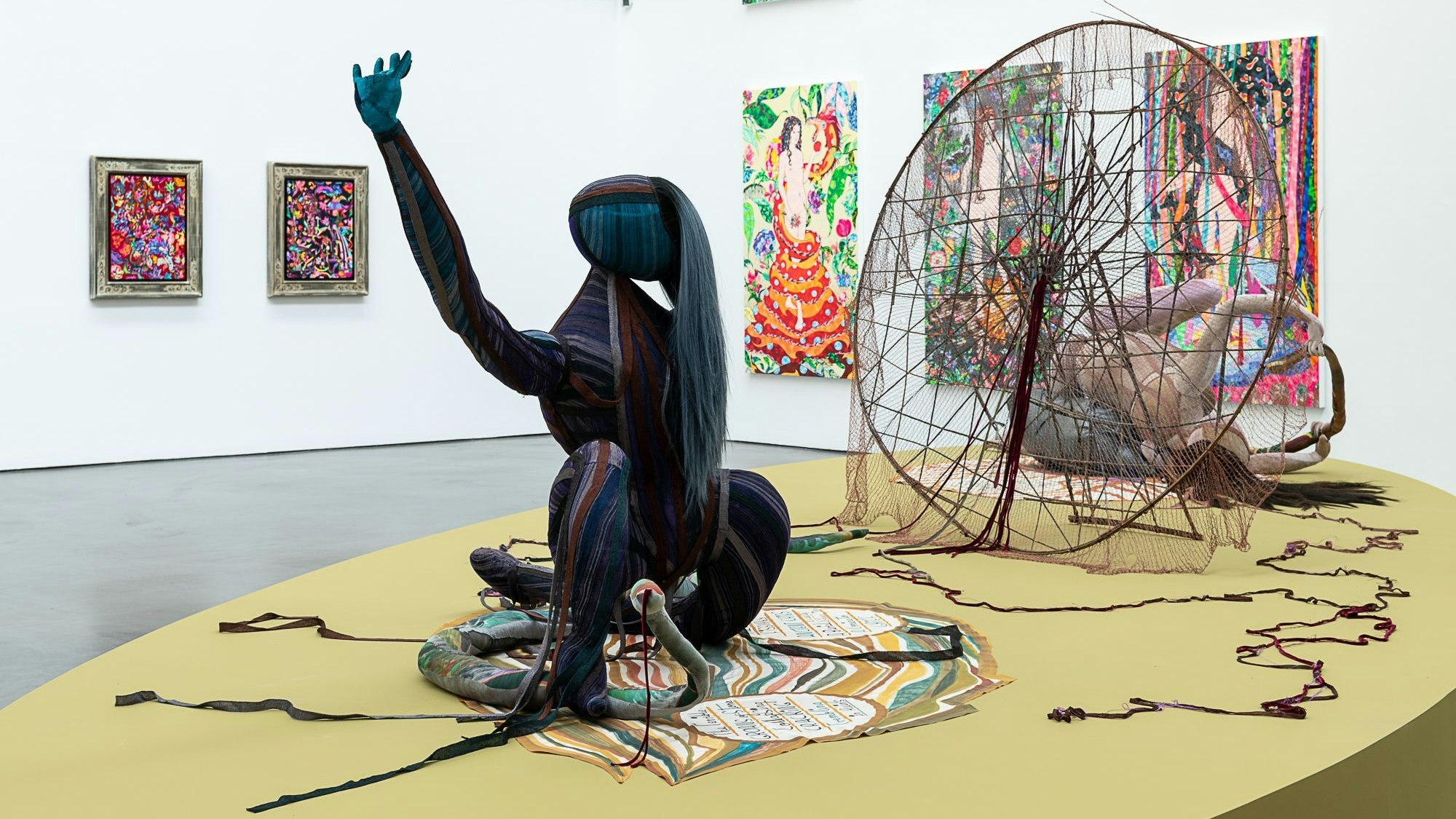 Im Vordergrund ist eine Skulptur auf einem Teppich, dahinter ein großes traumfängerartiges Netz und einige Bilder an den Wänden.