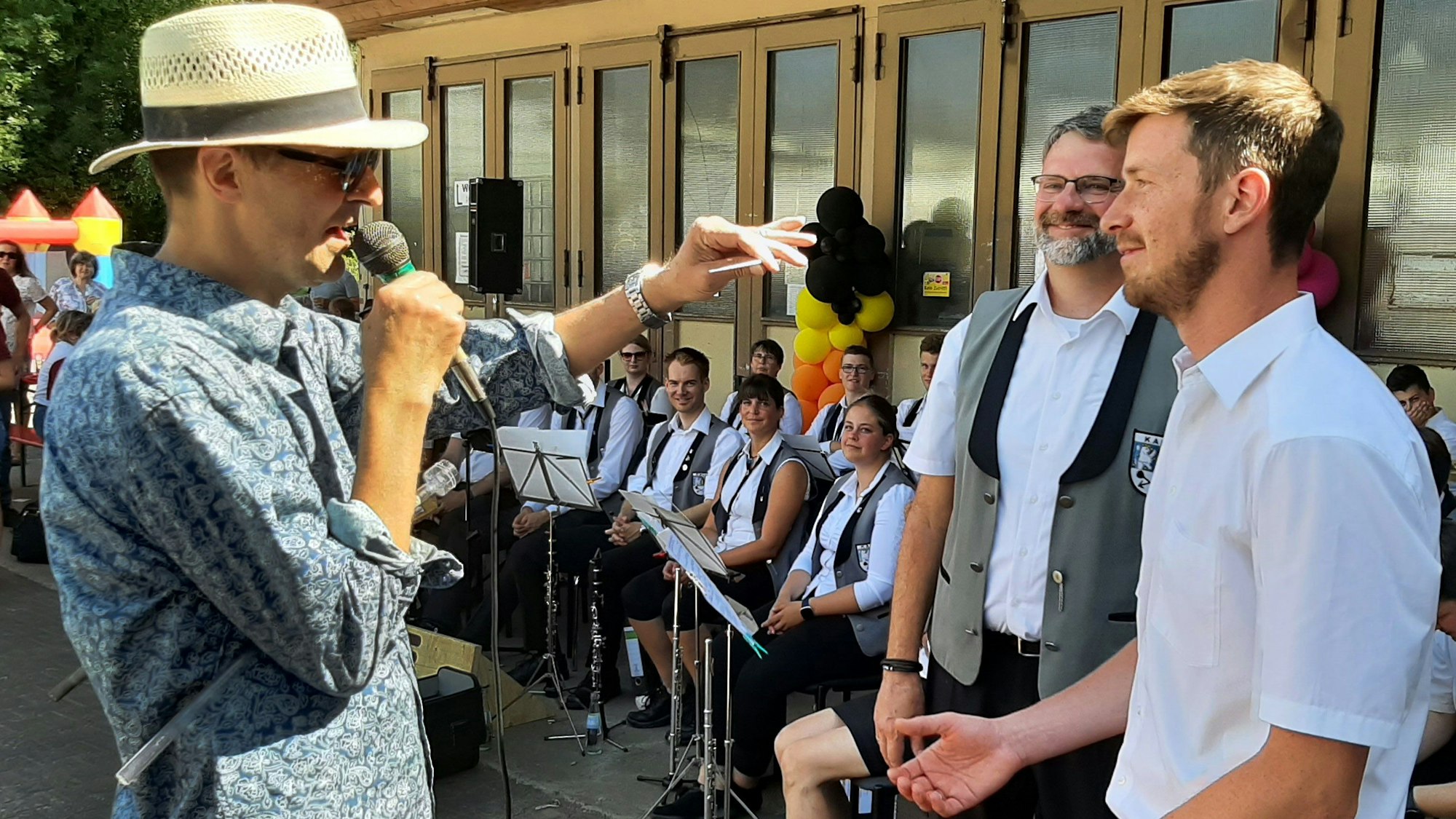 Ein Mann mit Strohhut übergibt den Taktstock an seinen Nachfolger.