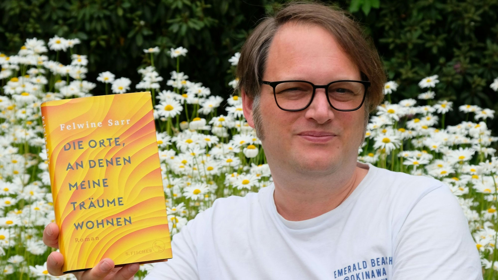 Gerrit Völker trägt eine Brille und ein weißes T-Shirt. Er hält Felwine Sarrs Buch mit einer Hand. Hinter ihm sind einige weiße Margeriten zu sehen.