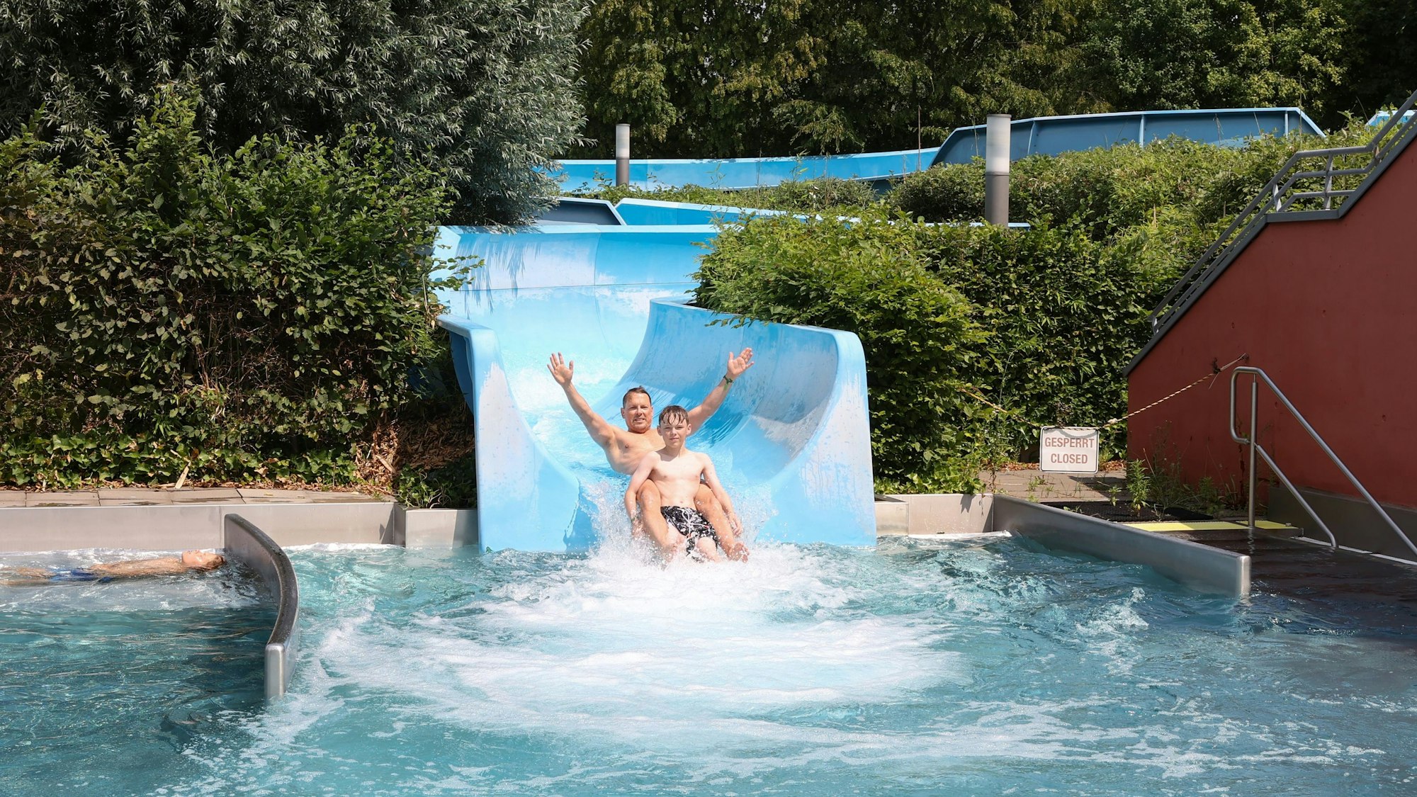 Ein Junge und sein Vater rutschen über eine halb-offene Rutsche ins Becken.