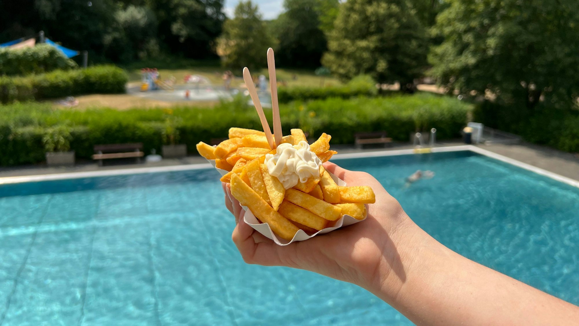 Eine Portion Pommes in einer Hand gehalten, dahinter sieht man ein Schwimmbecken.