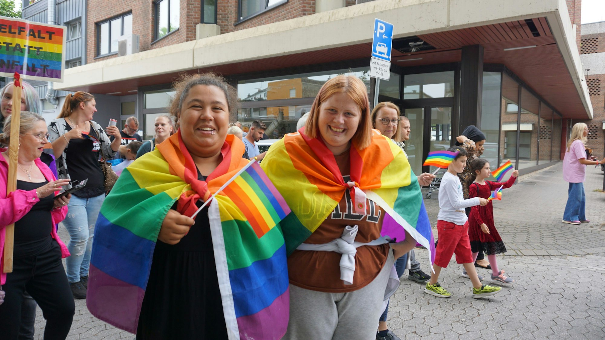 Zwei junge Frauen haben sich Regenbogenfahnen umgehangen. Eine von ihnen schwenkt zusätzlich eine Regenbogenflagge.
