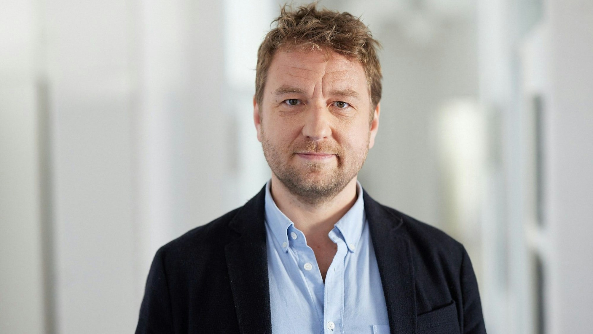 Tobias Bock trägt ein schwarzes Sakko und ein hellblaues Hemd. Er hat einen Drei-Tage-Bart.