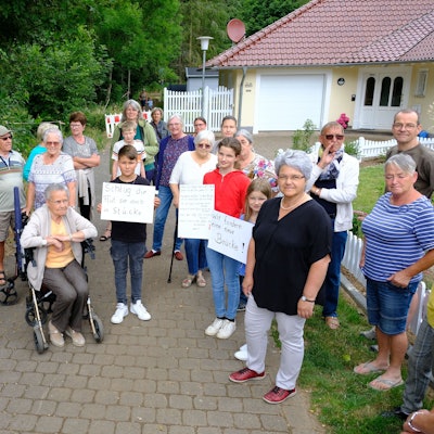 Eine Gruppe von Anliegern, darunter auch Kinder und eine Frau mit Rollator, hat sich auf einem Fußweg versammelt. Einige halten Plakate in die Kamera.