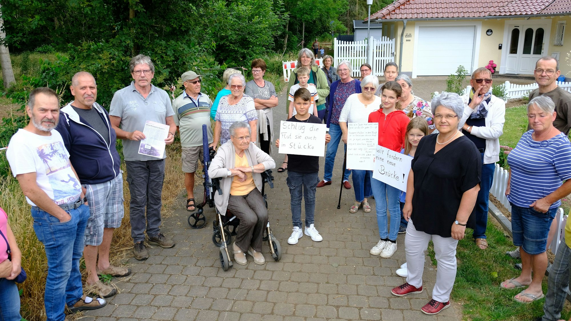 Eine Gruppe von Anliegern, darunter auch Kinder und eine Frau mit Rollator, hat sich auf einem Fußweg versammelt. Einige halten Plakate in die Kamera.