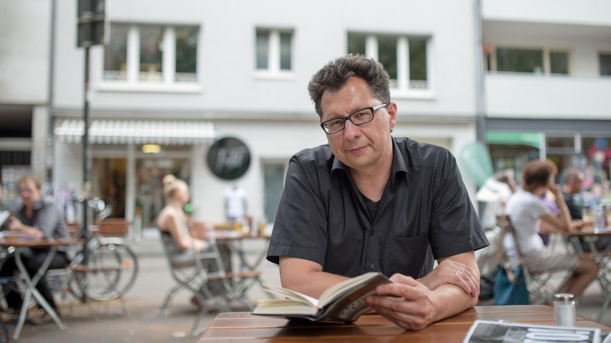 Uwe Kalkowski sitzt an einem Kaffee draußen und hat ein offenes Buch vor sich auf dem Tisch. Im Hintergrund sieht man verschwommen andere Gäste, eine Straße und dahinter eine Häuserreihe.