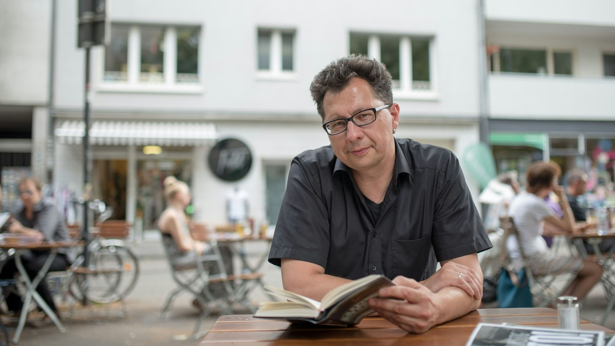 Uwe Kalkowski sitzt an einem Kaffee draußen und hat ein offenes Buch vor sich auf dem Tisch. Im Hintergrund sieht man verschwommen andere Gäste, eine Straße und dahinter eine Häuserreihe.