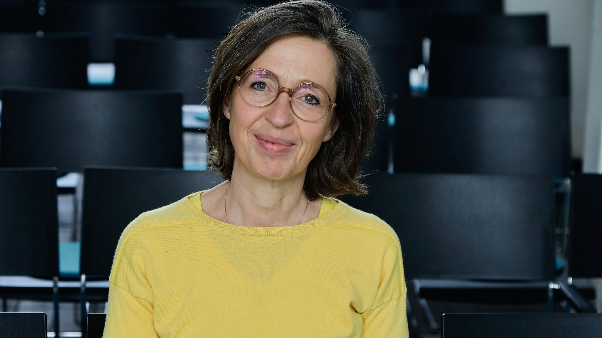 Bettina Fischer trägt eine Brille und einen gelben Pulli. Hinter und neben ihr sind schwarze Stühle.